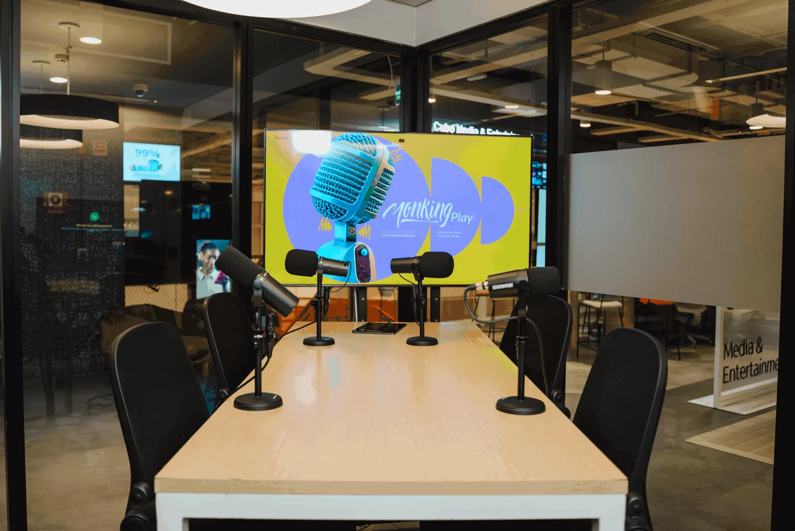 Estúdio do Cubo Itaú: mesa clara com quatro microfones, cadeiras e a TV do estúdio ao fundo, dentro do coworking